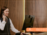 A woman in hotel reception uniform speaking on a telephone in front of a monitor, beside a wood-paneled wall. Social Comms UK asks, "What is the Best Phone System for Your Hotel?".