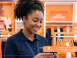 A smiling female retail worker in a navy blue polo shirt looking down at her smartphone inside a clothing store with orange signage.