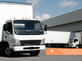 A white commercial delivery truck parked in the foreground of a large logistics warehouse facility, with a smaller white box truck parked at a loading bay in the background.