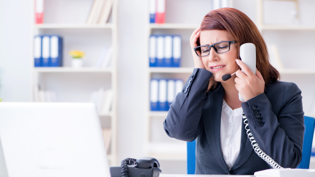 A frustrated female office worker wearing a headset and glasses, holding a traditional desk phone receiver to her ear while clutching her head in stress. She is sitting at a white desk with a laptop, appearing overwhelmed by technical issues or a poor connection.