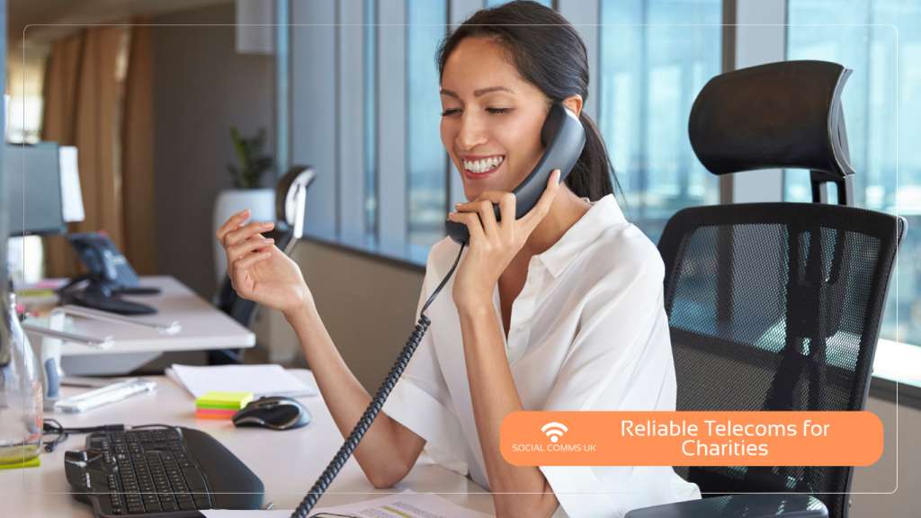 A smiling female office worker or helpline operator sitting at a desk and speaking on a telephone handset, featuring a Social Comms UK banner that reads 'Reliable Telecoms for Charities'.