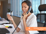 A smiling female office worker or helpline operator sitting at a desk and speaking on a telephone handset, featuring a Social Comms UK banner that reads 'Reliable Telecoms for Charities'.